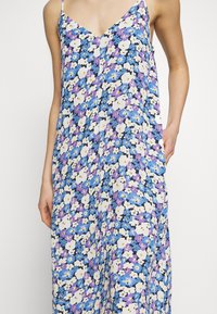 Kwiatowa sukienka z dekoltem w kształcie litery V, cienkimi ramiączkami i luźnym krojem. Tkanina przedstawia żywą mieszankę fioletowych, niebieskich i białych kwiatów na ciemnym tle.