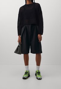 Pull noir en crochet à coupe ample, associé à un short noir, des chaussettes blanches et des chaussures de sport avec des accents verts. Porte un sac en filet noir.