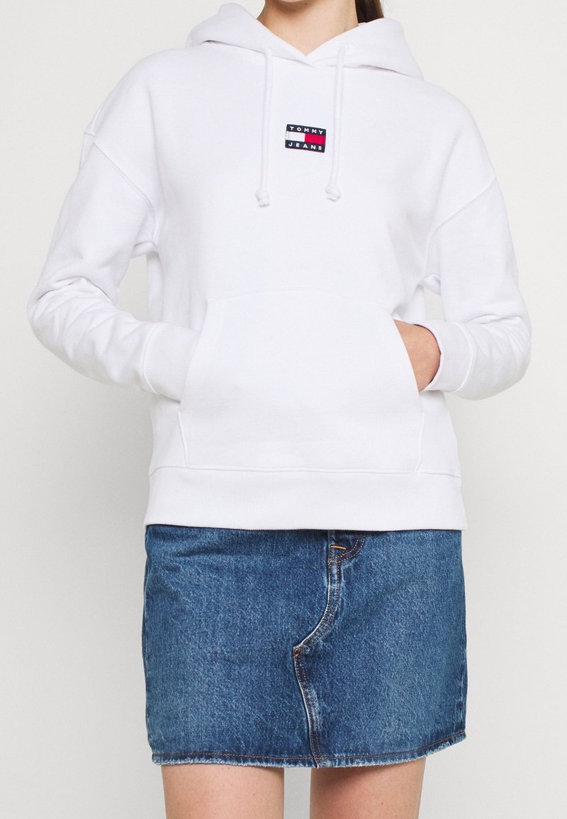 Sweat à capuche blanc avec poche avant et logo "Tommy Jeans", porté avec une jupe en jean bleue, les mains partiellement dans la poche du sweat.