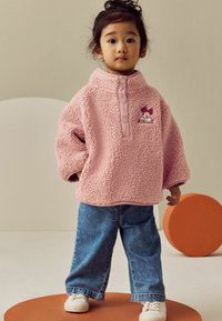 Rosa Fleece-Pullover mit hohem Kragen, Druckknöpfen und gesticktem Charakterdetail, kombiniert mit blauen Jeans im geraden Schnitt und weißen Sneakers.