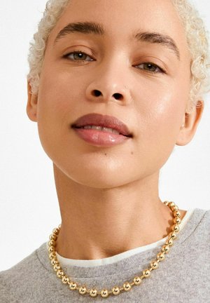 Ogrlica z zlatimi kroglicami, ki vsebuje okrogle, polirane kroglice, prikazana na svetlo sivi srajci z belim ovratnikom.