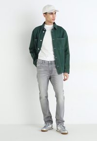 Πράσινο πουκάμισο με κουμπιά πάνω από λευκό μπλουζάκι, σε συνδυασμό με ανοιχτό γκρι τζιν και ασημί αθλητικά παπούτσια. Λευκό καπέλο φορεμένο στο κεφάλι.