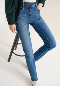 Personne portant un jean slim bleu, des baskets blanches et une veste boutonnée vert foncé, s'appuyant sur un tabouret noir.
