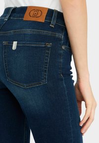 Jeans de mezclilla azul oscuro con cintura alta, con costuras amarillas, un parche de marca de cuero y un bolsillo trasero con un remache de metal.