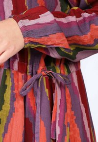 Mehrfarbiges Kleid mit einem Zickzackmuster in Rottönen, Rosa, Grün und Lila. Verfügt über elastische Bündchen und eine gebundenen Taille.