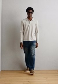 Lys beige langermet polo skjorte, blå denim bukser og brune joggesko. Modellen har på seg svarte solbriller og står mot en nøytral bakgrunn.