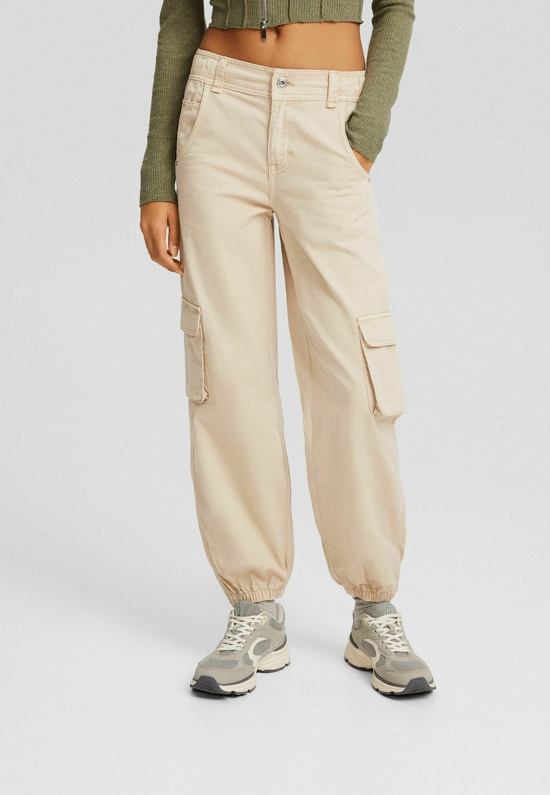 Pantalon cargo beige avec des poignets élastiques, porté avec des baskets grises et blanches et un top court vert à manches longues avec fermeture éclair.