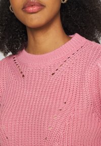 Pull en maille rose avec un col rond, présentant un motif texturé et de petits détails ajourés le long de l'épaule et du décolleté.