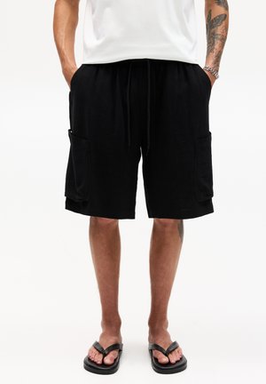 Shorts noirs en tissu doux, avec une taille élastique et un cordon de serrage, ainsi que deux poches latérales. Portés avec des tongs noires.