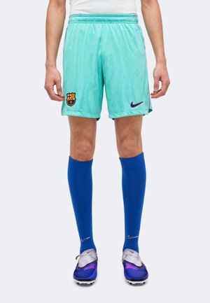 Homme portant un short de football vert clair du FC Barcelone, des chaussettes montantes bleues et des crampons violets et argentés, debout devant un fond uni.