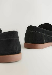 Chaussures slip-on en daim noir avec un bout rond, dotées d'une semelle en caoutchouc marron et de détails de couture contrastants sur le dessus.