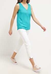 Turkusowa bezrękawnik z dekoltem w serek i zamkiem z przodu, zestawiony z białymi spodniami 3/4 z rozcięciami na dole oraz jasnoróżowymi sneakersami.