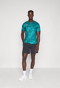 Homme portant une chemise à motifs teal à manches courtes, un short noir, des chaussettes blanches et des baskets blanches, se tenant devant un fond blanc uni.