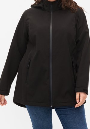 Giacca nera con cappuccio e maniche lunghe, con cerniera frontale e tasche con zip, indossata sopra jeans blu da una persona in piedi.