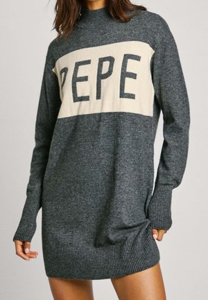 Vrouw die een lange grijze gebreide trui jurk draagt met een hoge hals en een crèmekleurige streep over de borst met letters "PEPE."