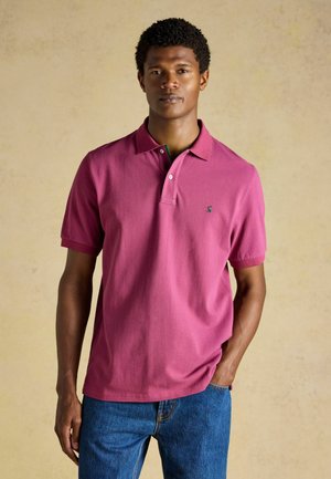 Uomo in piedi con una mano in tasca, che indossa una polo magenta a maniche corte e jeans blu su uno sfondo beige uniforme.
