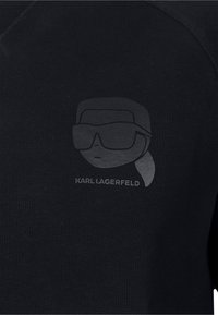 Sudadera negra con un diseño impreso gris de una figura con gafas de sol, acompañada del texto "KARL LAGERFELD." Textura suave.
