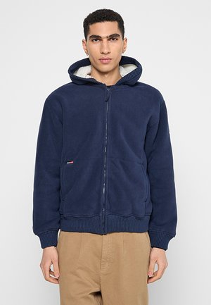 Ung mand iført en marineblå zip-up hættetrøje med fleeceforet hætte og beige bukser, stående mod en ensfarvet baggrund.