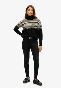 Schwarz-weißer gemusterter Pullover mit hohem Rollkragen, kombiniert mit schwarzen Skinny Jeans und schwarzen Schuhen. Der Pullover weist geometrische Muster auf.