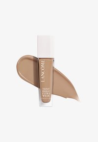 Fond de teint Lancôme Teint Idole Ultra Wear dans un flacon en verre transparent avec un bouchon blanc, à côté d'un échantillon beige clair. Texture lisse.