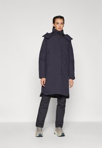 Cappotto lungo blu scuro con colletto alto, chiusura zip e cappuccio regolabile; abbinato a pantaloni coordinati e sneakers grigio chiaro.