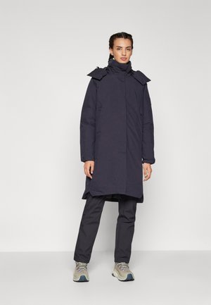 Donkerblauwe lange jas met hoge kraag, ritssluiting en verstelbare capuchon; gecombineerd met bijpassende broek en lichtgrijze sneakers.