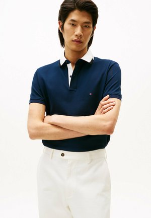 Polo shirt blu navy con colletto bianco a contrasto, maniche corte e un piccolo logo sul petto; abbinato a pantaloni bianchi.