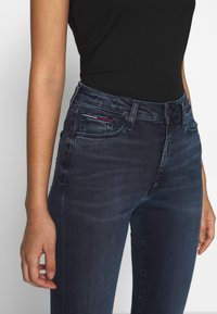 Jeans de mezclilla azul oscuro con un corte slim, que presentan un diseño estándar de cinco bolsillos y detalles de costura sutiles. La tela tiene una textura suave.