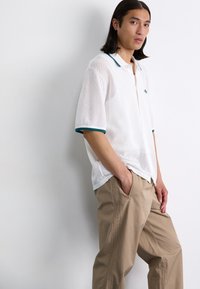 White mesh polo shirt with teal accents on the collar and sleeves, paired with beige pants featuring side zippers and stitching details.