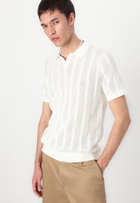 Camisa pólo branca de manga curta com um padrão de malha texturizada, apresentando riscas verticais e colarinho e punhos canelados.