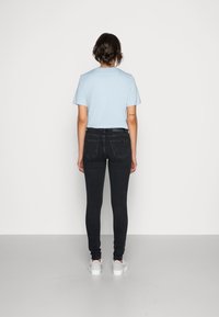 Person med kort hår iklädd en ljusblå t-shirt, svarta tajta jeans och vita sneakers, stående vänd mot en enkel vit vägg.