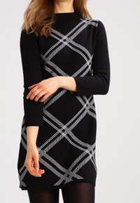 Czarna sukienka z białym, ukośnym paskowanym wzorem, długimi rękawami, wysokim dekoltem i lekko rozkloszowanym dołem, wykonana z miękkiego materiału.