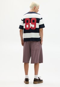 Striped navy and white t-shirt with large red number 89 on back; paired with loose purple shorts and brown shoes. Back view.