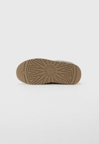 Sole of a tan UGG slipper with textured tread pattern and "UGG" logo at the center, displayed against a white background.