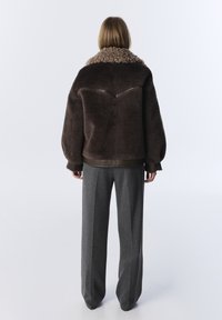 Personne debout avec le dos visible, portant une veste en shearling brun foncé avec un col duveteux et un pantalon large gris.