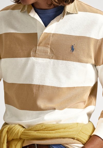 Vīrietis, kurš valkā bēša un baltas svītras ragbija kreklu ar zilu apakškreklu, dzeltens džemperis sasiets ap vidukli, un daļēji redzami gari cirtaini mati.