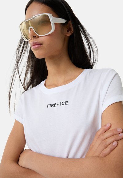 Hvid bomulds t-shirt med rullede ærmer, der prydes af teksten "FIRE+ICE" trykt i fed sort. Store hvide solbriller med lyse beige linser.