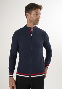 Marineblaue Zip-Up-Strickjacke aus strukturiertem Material, mit rot-weißen gestreiften Akzenten an den Bündchen und am Saum. Mit Stehkragen und taillierter Passform.