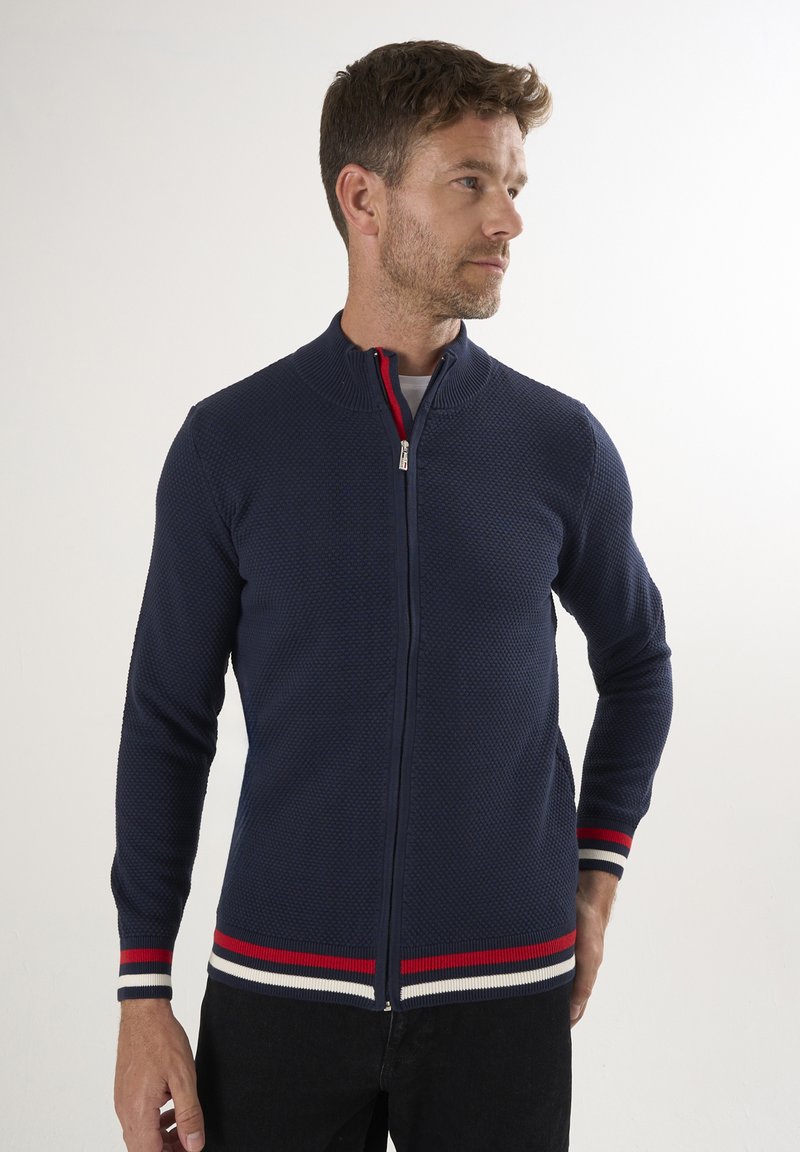 Marineblaue Zip-Up-Strickjacke aus strukturiertem Material, mit rot-weißen gestreiften Akzenten an den Bündchen und am Saum. Mit Stehkragen und taillierter Passform.