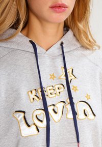 Sudadera gris con cordones azul marino, que presenta el texto bordado "KEEP LOVIN" y estrellas en acentos amarillos y negros. Textura de tela suave.