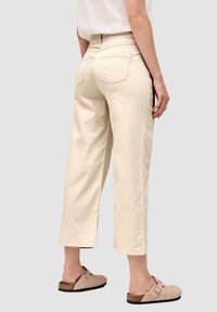 Pantalones cortos beige con dos bolsillos traseros, diseño de pierna recta y material de algodón suave. Llevado con calzado beige tipo slip-on.