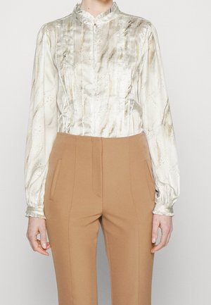Personne portant un chemisier en soie blanche avec des accents dorés et un pantalon camel taille haute, debout devant un fond uni.