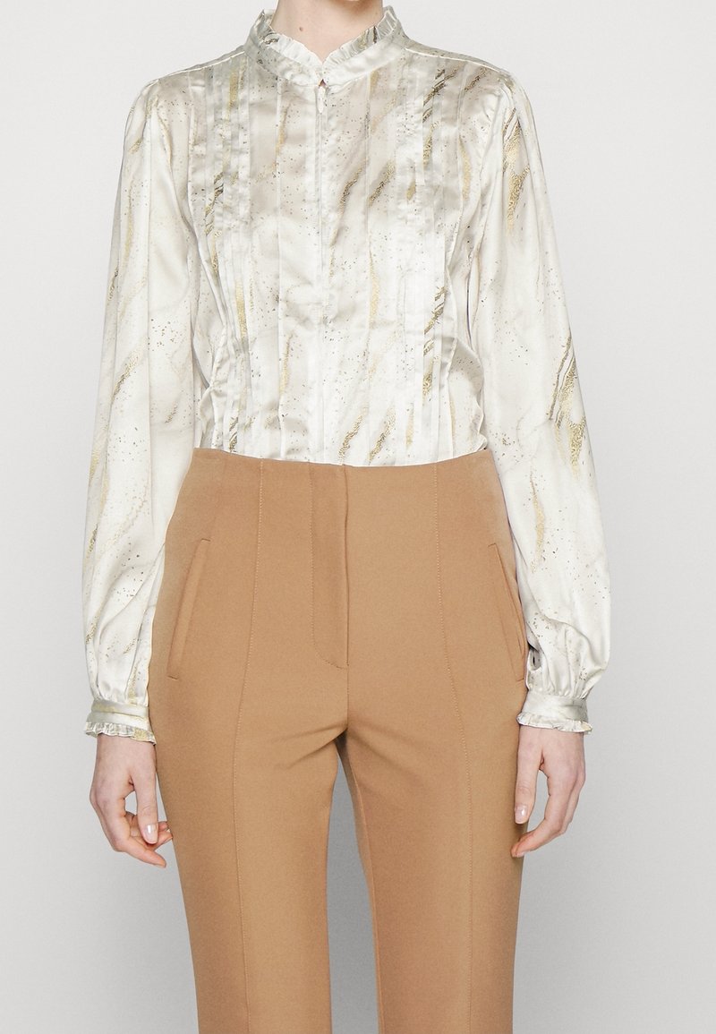 Personne portant un chemisier en soie blanche avec des accents dorés et un pantalon camel taille haute, debout devant un fond uni.