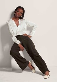 Weiße Satinbluse mit V-Ausschnitt und langen Ärmeln, kombiniert mit braunen, hoch geschnittenen, weiten Hosen und weißen Blockabsatzschuhen.