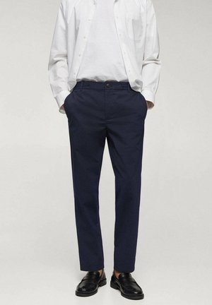 Mann trägt dunkelblaue Anzughose, schwarze Loafers, weißes T-Shirt und weißes Hemd mit Knöpfen mit Händen in den Taschen.