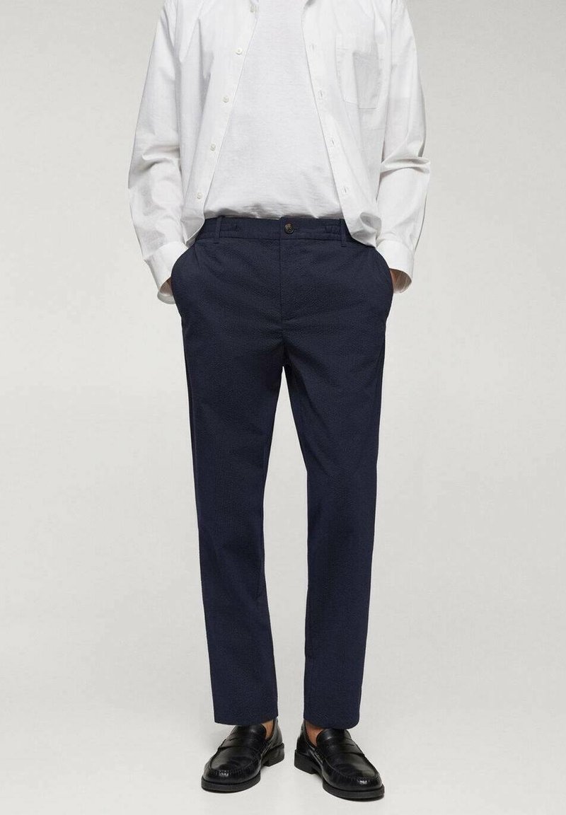 Homme portant un pantalon habillé bleu marine, des mocassins noirs, un t-shirt blanc et une chemise blanche boutonnée, les mains dans les poches.