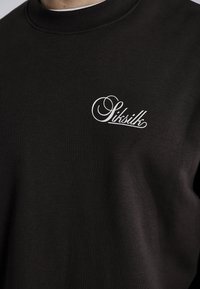 Czarny sweterek z bawełny z ściągaczem. Posiada biały haftowany logotyp na piersi. Gładka tekstura, luźny krój.