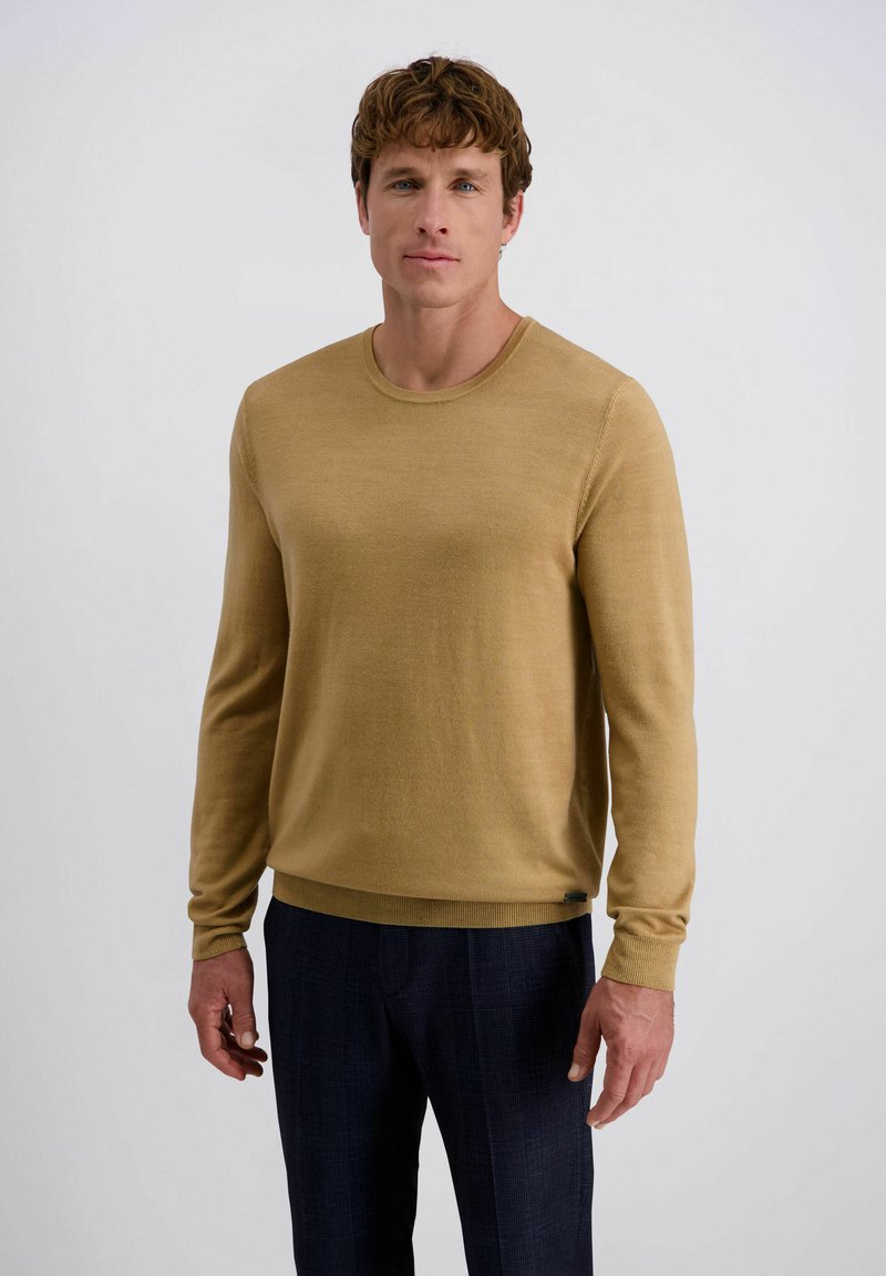 Mann mit kurzen lockigen Haaren, der einen senfgelben Pullover und eine dunkle Marinehose trägt, vor einem einfachen weißen Hintergrund.