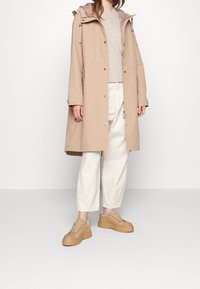 Beiger wasserdichter Mantel mit Kapuze, zwei Fronttaschen, beiges T-Shirt, helle Hose und beige klobige Sneakers. Glatte Textur, lässiges Design.