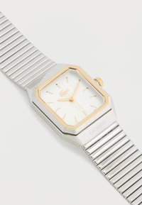 Ασημένιο μεταλλικό ρολόι χειρός με ορθογώνιο καντράν με χρυσές λεπτομέρειες, δείκτες και αριθμούς σε χρυσό, και εύκαμπτο λουράκι με ραβδώσεις.